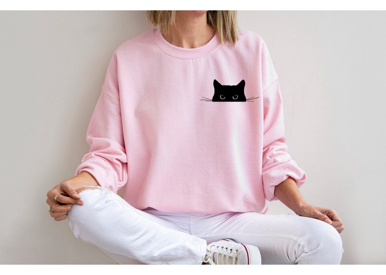 May include: A pink sweatshirt with a black cat peeking out from the pocket. The cat has large eyes and a small nose.