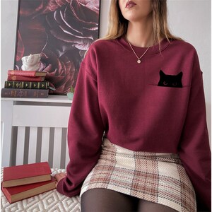 May include: A maroon sweatshirt with a black cat peeking out of a pocket. The sweatshirt has a crew neck and long sleeves.