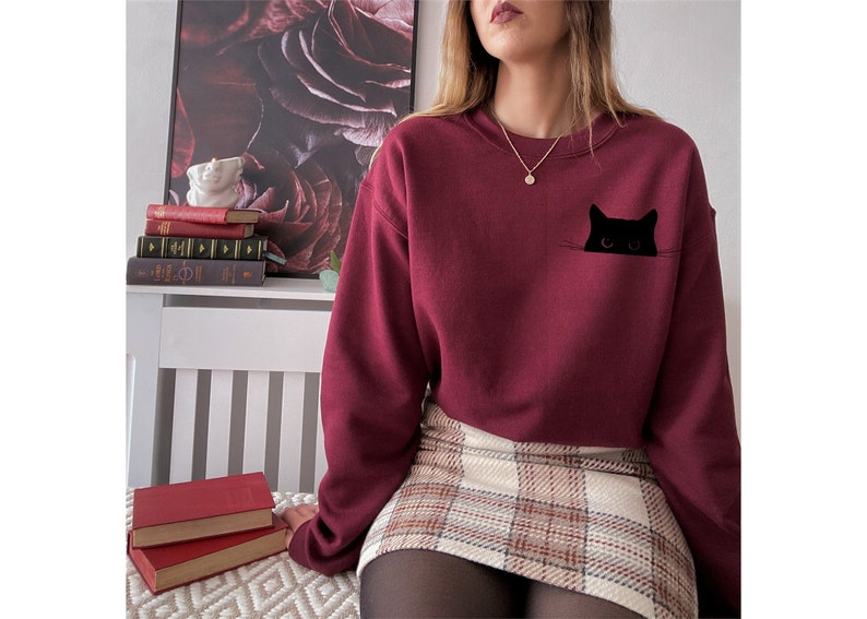 May include: A maroon sweatshirt with a black cat peeking out of a pocket. The sweatshirt has a crew neck and long sleeves.