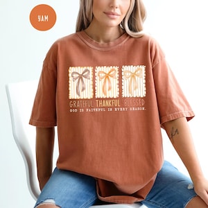 May include: A rust-colored t-shirt featuring a graphic with three bow designs in brown, orange, and yellow. The text reads "Grateful Thankful Blessed" and "God is faithful in every season."