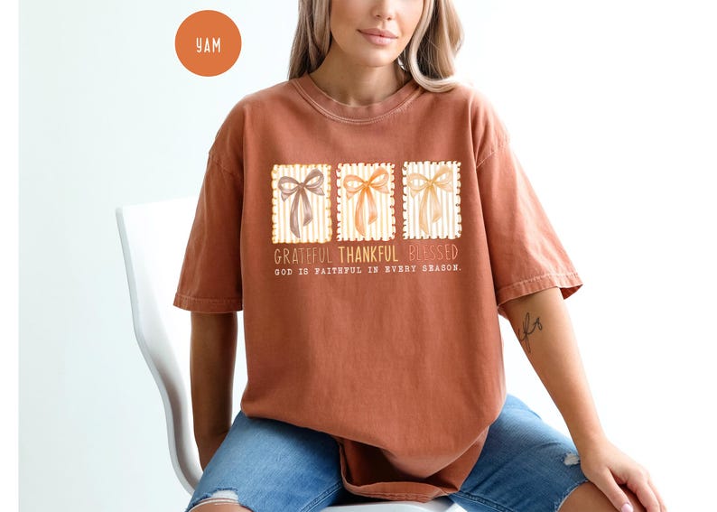 May include: A rust-colored t-shirt featuring a graphic with three bow designs in brown, orange, and yellow. The text reads "Grateful Thankful Blessed" and "God is faithful in every season."