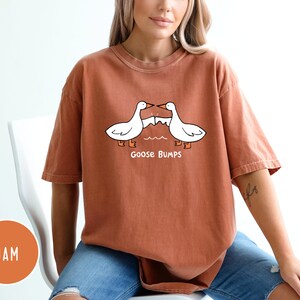 Może przedstawiać: Rdzawa koszulka z kresk&oacute;wkowym wzorem dw&oacute;ch białych gęsi zwr&oacute;conych do siebie, z napisem "Goose Bumps" poniżej. Gęsi są narysowane czarnymi konturami i pomarańczowymi stopami. Koszulka ma kr&oacute;tkie rękawy i luźny kr&oacute;j.