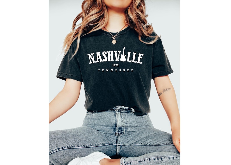 May include: Black t-shirt with the word "NASHVILLE" in white, a guitar graphic, and the text "1972 TENNESSEE". The shirt is worn with blue jeans.