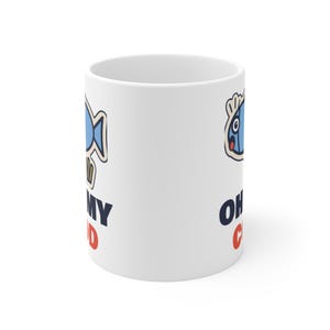 May include: A white ceramic mug featuring a cartoon blue fish graphic and the text "OH MY COD" printed in dark blue and red. The design includes a stylised fish with a surprised expression, positioned above the bold, multi-coloured lettering.