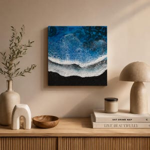 Op de afbeelding: Vierkante wandkunst die een kustlandschap toont met een zwart zandstrand en oceaangolven in blauw en wit. Het kunstwerk is gemonteerd op een neutraal gekleurde muur, met decoratieve items op een houten oppervlak eronder.