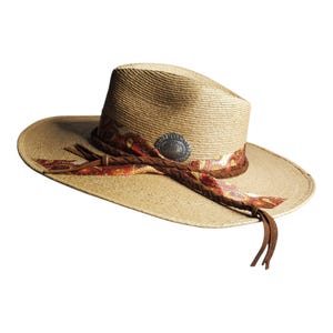 May include: A tan straw hat with a wide brim and a pinched crown. The hat features a decorative band with a red, orange, and yellow paisley pattern, a braided brown leather cord, and a circular silver-toned metal concho accent on the front.