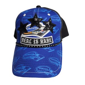 May include: A blue and black baseball cap featuring a repeating Samsung 500 logo pattern. The front displays three black sequined stars, a Samsung 500 Texas Motor Speedway patch, and a white rectangular patch with the text REAL IS RARE.