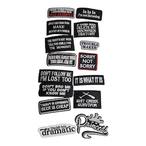 May include: A collection of black and white embroidered patches with various phrases and designs. The patches include text like "Bad Decisions Make Good Stories," "It Is What It Is," and "Shit Creek Survivor." Some patches have decorative elements.