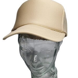 May include: Tan trucker hat with a mesh back and a curved brim. The front panel is solid tan, and the brim is trimmed with a thin, tan rope. The hat is displayed on a clear glass mannequin head.