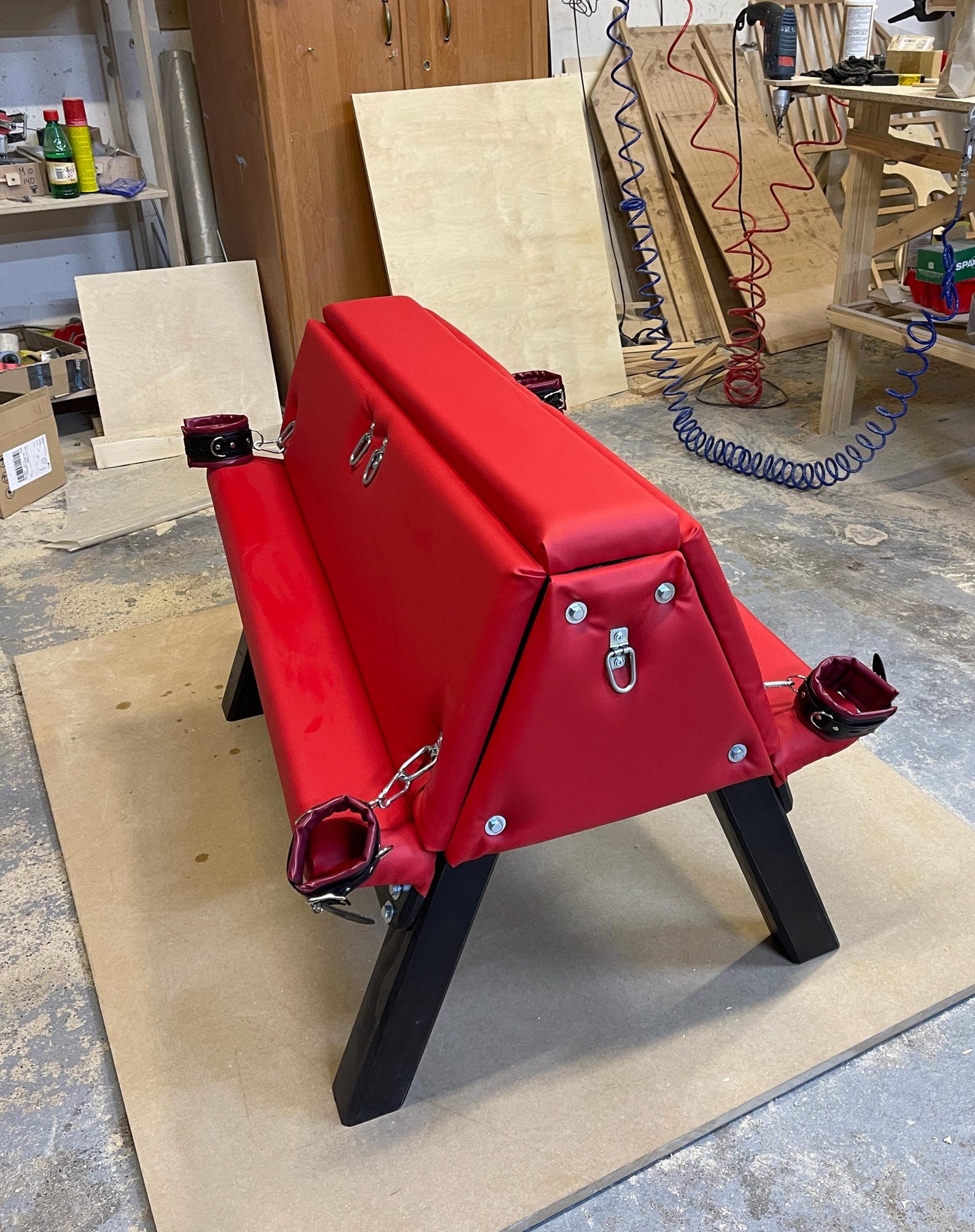 Red padded bdsm bench with black legs and attached cuffs, ideal bondage furniture for sex dungeon or bdsm room setups.