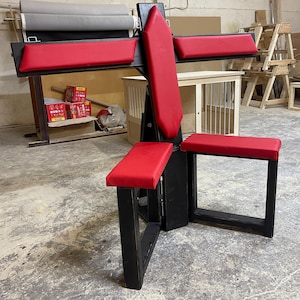 May include: A black wooden structure with red padded seating. The design features a central vertical support with horizontal armrests and a lower seat, all with red upholstery. The piece is designed for a specific purpose.