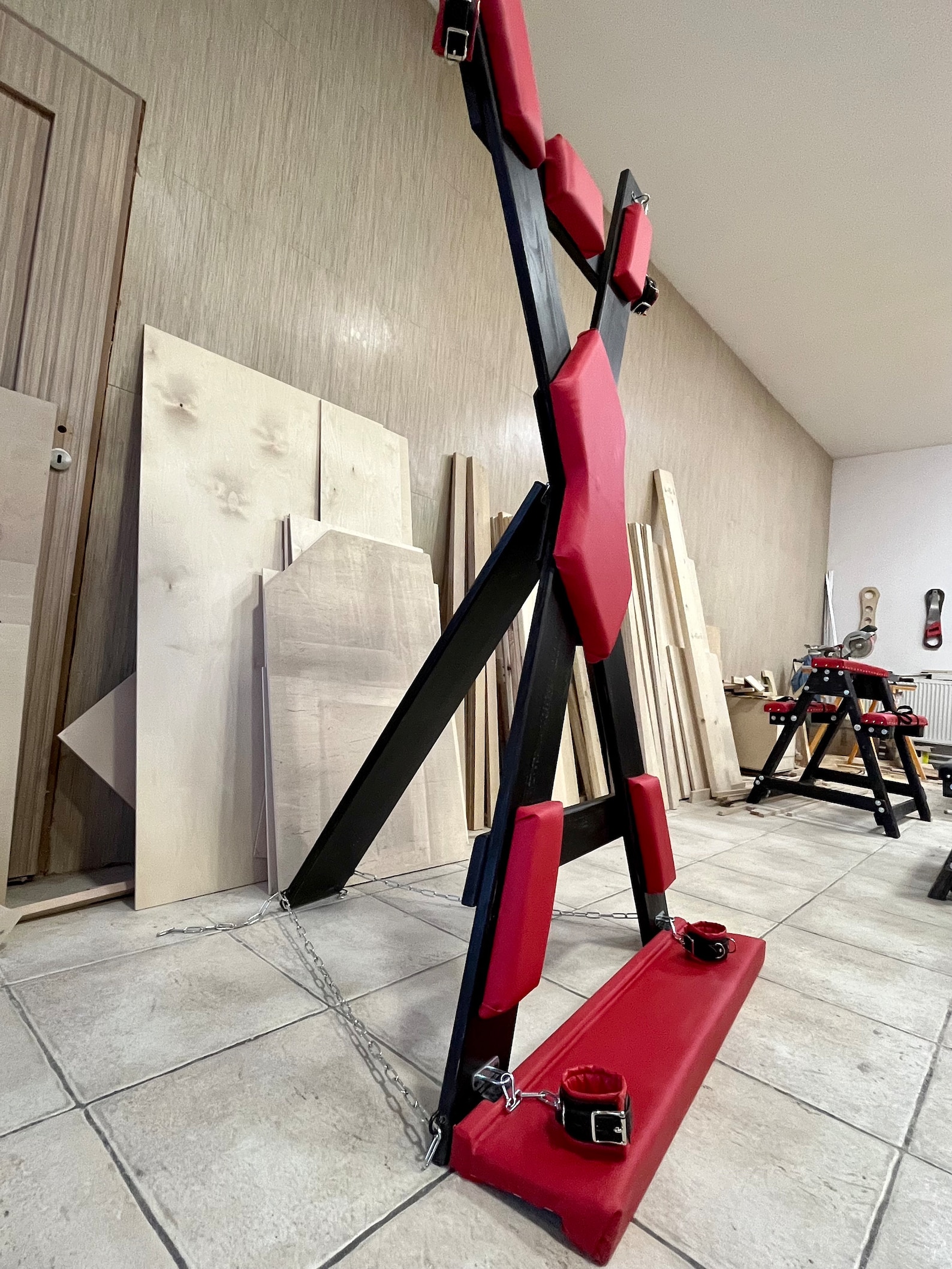 Angled view of bondage furniture St. Andrew’s Cross with footrest and restraints for sex dungeon or BDSM room setup.