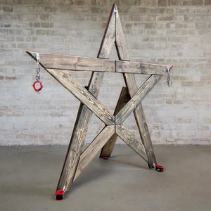 Könnte beinhalten: Eine sternförmige Holzstruktur mit roten Metallringen, die an Ketten befestigt sind. Die Struktur ist aus grob behautem Holz gefertigt und hat ein rustikales Aussehen.