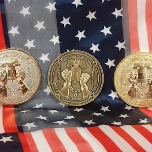 May include: Three gold-colored commemorative coins with a design of a bell and shackles. The coins are on a red, white, and blue American flag. The text on the coins reads "Juneteenth Day, June 19, 1865", "Freedom", "Emancipation Proclamation", "January 1, 1863", and "Liberty".