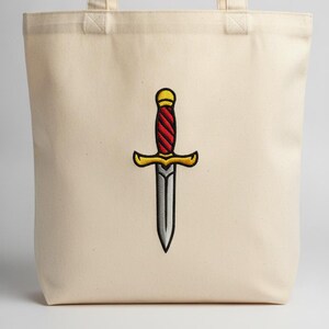 Puede incluir: Un bolso tote de lona color crema con un diseño bordado centrado de una daga. La daga tiene una hoja plateada, una guarda y un pomo dorados, y un mango con un patrón de espiral rojo y dorado delineado con hilo de color negro.