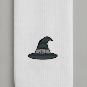 Può includere: Un canovaccio da cucina bianco con trama a nido d'ape che presenta un disegno centrale ricamato a macchina raffigurante un cappello da strega nero con una fascia grigio chiaro e una fibbia quadrata. Il tessuto ha una texture a griglia.