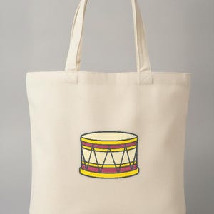 May include: A cream-colored canvas tote bag featuring a centered graphic illustration of a drum. The drum is depicted with yellow rims, a deep red horizontal band, and black tension rods arranged in a triangular pattern against a cream body.