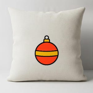 May include: A square off-white throw pillow featuring a centered graphic of a round red Christmas ornament. The ornament has a horizontal yellow stripe across the middle and a yellow cap at the top, all outlined in a bold black line.