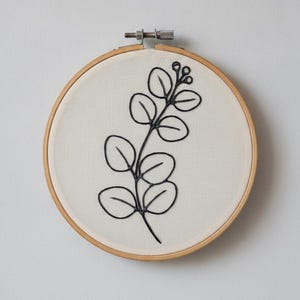 May include: A circular wooden embroidery hoop containing off-white fabric with a black thread outline of a eucalyptus branch. The branch features several rounded leaves and small buds at the top, secured by a metal tension screw at the top.