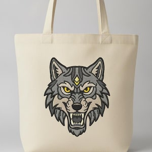 May include: A cream-colored canvas tote bag featuring a centered, detailed embroidery of a snarling wolf head. The wolf design uses shades of gray, black, and white with piercing yellow eyes and a small yellow diamond-shaped accent on its forehead.