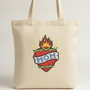 Puede incluir: Un bolso de tela de color natural con un diseño bordado central de un corazón rojo envuelto en espinas verdes, coronado con llamas amarillas y naranjas, y una banda azul con la palabra MOM bordada en blanco en el centro.