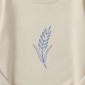 May include: A cream-colored crewneck sweatshirt featuring a centered, minimalist blue line art illustration of a single wheat stalk. The garment has long sleeves, a ribbed collar, and a soft, smooth fabric texture throughout the front panel.