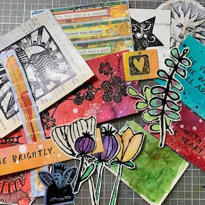 May include: A collection of mixed media paper ephemera including hand-drawn floral cutouts, stamped sun motifs, and rectangular cards with text such as SHINE BRIGHTLY, BE YOU, THE WORLD WILL ADJUST, and LIVE WITH ALL YOUR HEART in various colors.