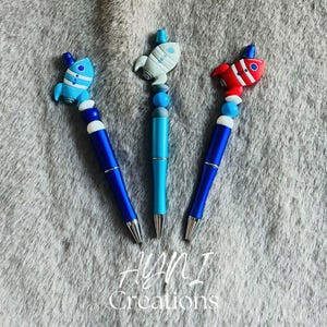 Pode incluir: Três canetas com tampas de foguete azuis, cinzentas e vermelhas. As canetas têm um corpo azul com detalhes prateados e um design de contas. A imagem também inclui o texto "AYNI Creations" em branco.