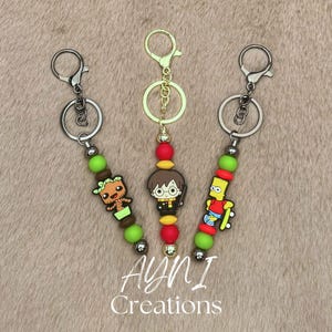 Peut inclure: Trois porte-clés avec des breloques de personnages. L'un représente Groot, un autre Harry Potter et le troisième Bart Simpson. Chaque porte-clés a un fermoir en métal, des perles vertes, rouges et argentées, et le texte "AYNI Creations".