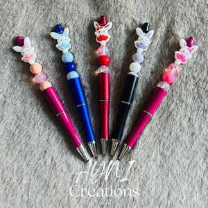 Pode incluir: Cinco canetas coloridas com enfeites de coelho. As canetas têm corpos rosa, azuis, vermelhos e pretos. Cada caneta apresenta um design de coelho diferente com uma cartola e uma fileira de contas. O texto "AYNI Creations" está na parte inferior.