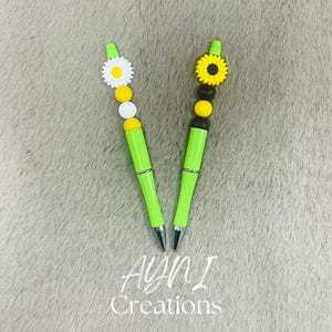 Könnte beinhalten: Zwei grüne Stifte mit dekorativen Aufsätzen. Ein Stift hat eine weiße Gänseblümchen mit gelbem Zentrum, gelben und weißen Perlen. Der andere Stift hat eine gelbe Sonnenblume mit braunem Zentrum, gelben Perlen. Der Text "AYNI Creations" steht unten.