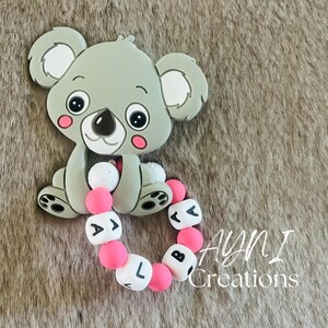 Pode incluir: Um mordedor de silicone cinza em forma de coala com contas rosa e brancas que soletram "A.B.L." O mordedor está sobre uma superfície marrom e felpuda.  AYNI Creations está escrito em texto branco sobre fundo preto.