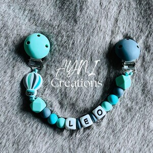 May include: A pacifier clip featuring a mint green and dark gray color scheme with silicone beads, a hot air balloon charm, and white letter beads spelling LEO. The clip includes two circular fasteners at each end of the beaded cord.