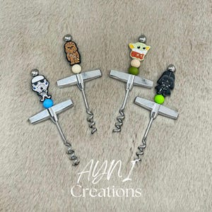 Op de afbeelding: Vier zilveren kurkentrekkers met kralen met personagethema. De kralen tonen Chewbacca, Baby Yoda, Darth Vader en een Stormtrooper-schedel. De tekst "AYNI Creations" is zichtbaar in witte letters.
