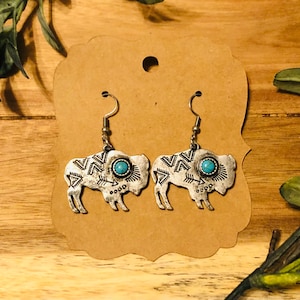 May include: Silver dangle earrings featuring a buffalo silhouette with turquoise accents and geometric patterns.