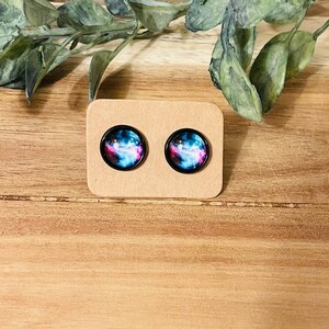 May include: A pair of galaxy-themed stud earrings with a black background and pink, blue, and white nebulae. The earrings are displayed on a brown card.