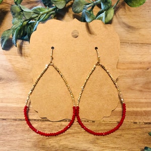 May include: A pair of gold-colored teardrop earrings with a red beaded bottom. The earrings are displayed on a brown card with a small hole at the top. The background is a wooden surface with green foliage.