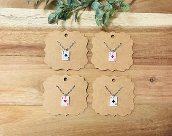 Puede incluir: Cuatro collares con dijes de palo de cartas sobre cartulina marrón. Los dijes son un as de picas, un as de corazones, un as de diamantes y un as de tréboles.
