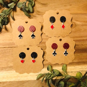 May include: Three pairs of earrings with glitter and card suit charms. The earrings are displayed on brown card stock. The charms are a spade, a club, and a diamond.