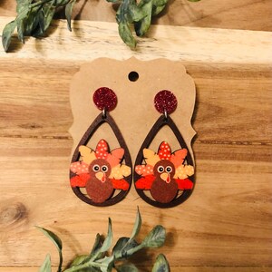 May include: A pair of brown wooden dangle earrings with a turkey design. The turkey is brown, orange, and red with white polka dots. The earrings have red glitter accents.