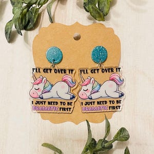 May include: A pair of clear acrylic earrings featuring a unicorn with the text "I'll get over it, I just need to be dramatic first". The unicorn is lying down with its eyes closed. The earrings have a teal glitter accent.