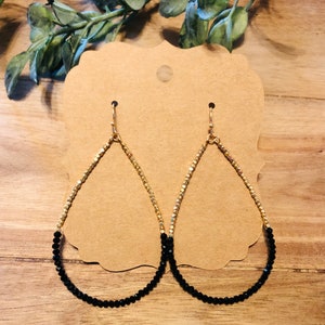 May include: A pair of teardrop-shaped earrings with gold and black beads. The earrings are made of small, round beads that are strung together to form a teardrop shape. The top half of each earring is made of gold beads, while the bottom half is made of black beads.