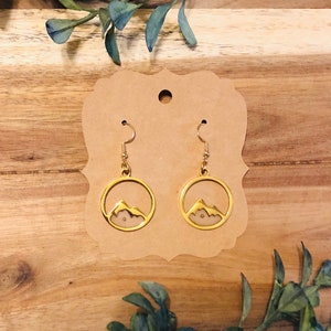 May include: Gold earrings with a mountain silhouette design. The earrings are on a brown card.