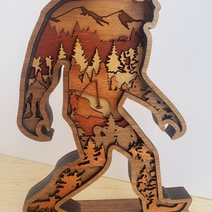 May include: A wooden sculpture of Bigfoot walking through a forest. The sculpture is made of multiple layers of wood, creating a 3D effect. The forest scene includes trees, mountains, and a small lake.