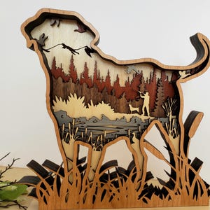 May include: A wooden cutout of a dog standing in a field with a hunter and a dog in the background. The dog is made of multiple pieces of wood, creating a layered effect. The hunter and dog are silhouetted in the background.