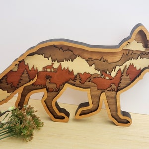 Puede incluir: Una silueta de zorro de madera con un diseño en capas que presenta una escena de bosque en su interior. El zorro es marrón y la escena del bosque está hecha de diferentes tonos de madera marrón y beige.