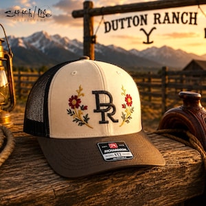 May include: A cream-colored trucker hat with a black mesh back and a dark brown brim. The front features an embroidered black DR monogram flanked by two floral stems with red and yellow flowers. A Richardson 112 sticker is on the brim.