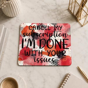 Puede incluir: Una alfombrilla de ratón rectangular con el texto "CANCEL MY subscription I'M DONE WITH YOUR issues" en negro sobre un diseño abstracto rojo y blanco. La alfombrilla está sobre una superficie de mármol, con una taza de café y un bloc de notas cerca.