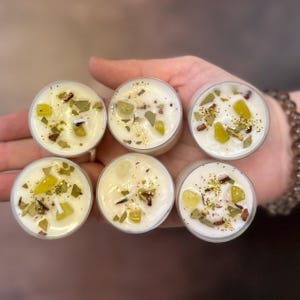 Peut inclure: Six petites bougies chauffe-plat en verre transparent avec de la cire blanche et des paillettes vertes et dorées incrustées. Les bougies sont disposées en cercle sur une main.