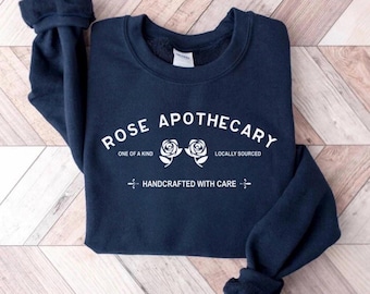 Puede incluir: Una sudadera azul marino con el texto blanco "ROSE APOTHECARY" y dos ilustraciones de rosas. La sudadera también muestra las frases "ONE OF A KIND", "LOCALLY SOURCED" y "HANDCRAFTED WITH CARE" en letras blancas.